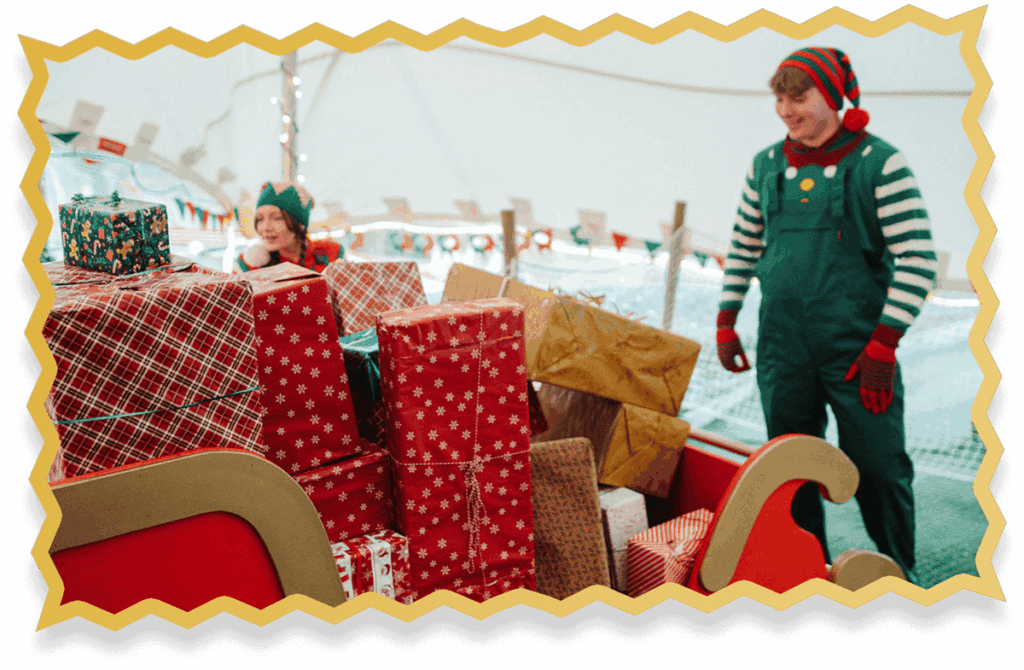 Two people dressed as Christmas elves stand by a large sleigh filled with wrapped holiday presents at Jingle Bell Junction. The festive scene sparkles with decorations and lights in the background.