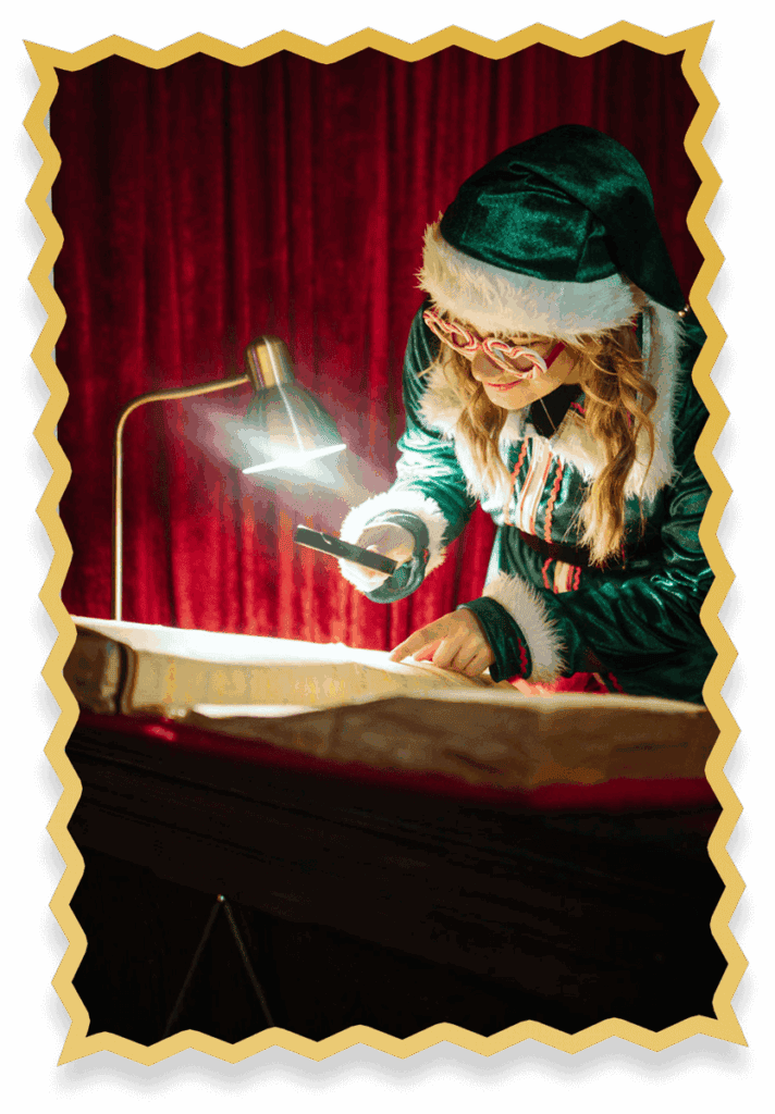 A person dressed as an elf in a green costume with white fur trim reads a large book under a desk lamp at Jingle Bell Junction, smiling and holding a magnifying glass, against a backdrop of red curtains.