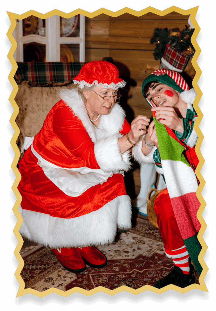 Mrs Claus, dressed in red and white, helps a smiling elf in a festive costume with a colourful scarf inside a cosy, decorated holiday room.