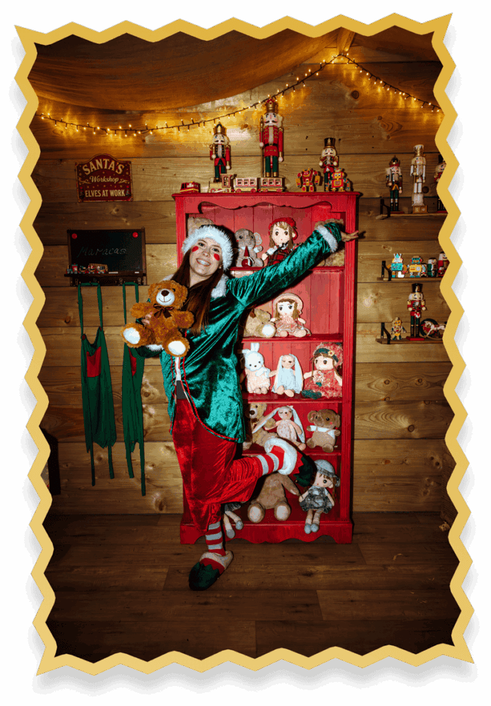 A person dressed as an elf poses playfully in front of a red shelf filled with soft toys and dolls. The wooden room is decorated with lights and holiday figurines, creating a festive, cheerful atmosphere.