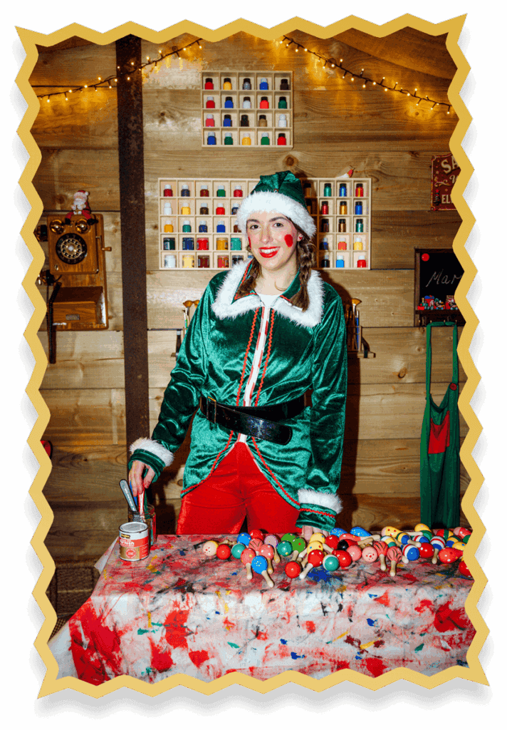 A person dressed as a cheerful Christmas elf stands at a colourful, paint-splattered table filled with painted wooden balls, inside a festively decorated wooden room with fairy lights.