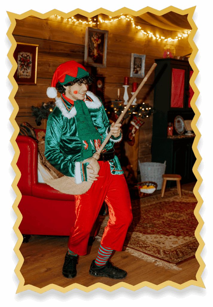 A person dressed as an elf in a festive room strums a broom like a guitar, smiling and wearing a green velvet jacket, red trousers, elf hat, and striped socks. Holiday decorations are visible in the background.